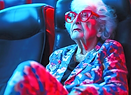 An elderly woman with white hair and red glasses sits alone in a movie theater, illuminated by the red and blue light.