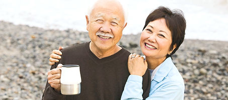 An elderly Asian couple is standing closely together on a rocky beach, smiling at the camera. The man is wearing a brown jacket and holding a silver travel mug. The woman is wearing a blue windbreaker and has her arm around the man's shoulder. The background shows the ocean and a large rock wall.