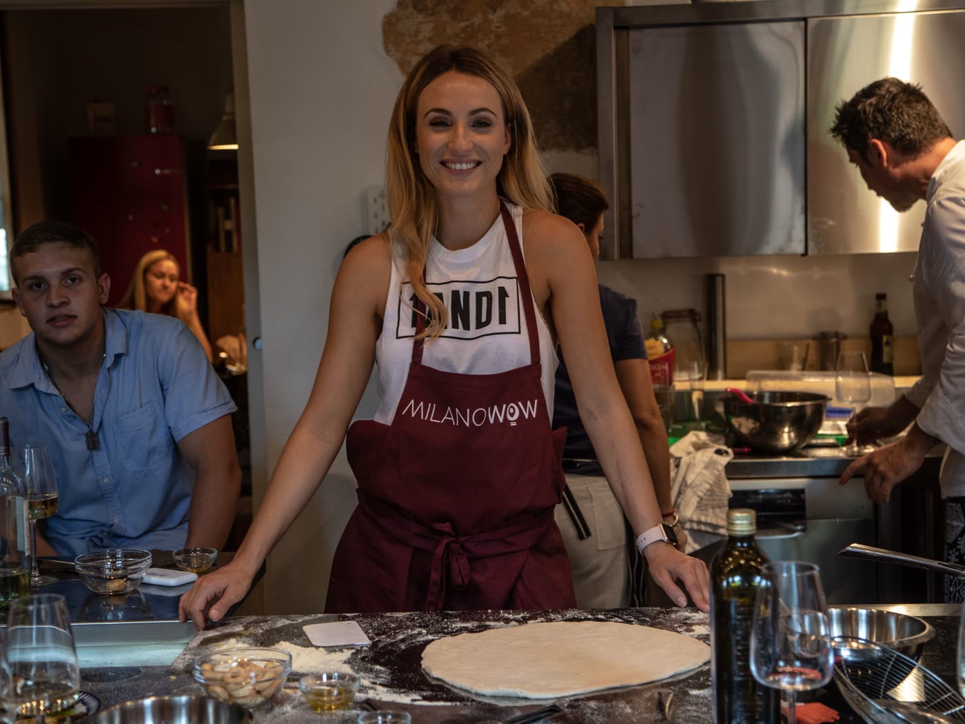 A participant in a branded apron at the counter during the Milan culinary session, the chef working behind