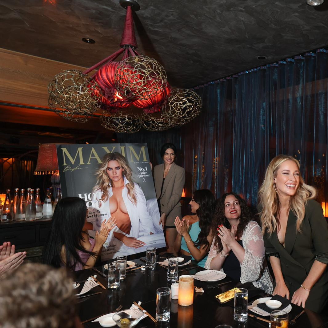 Guests applauding around the candlelit dinner table at the Maxim Hot 100 cover dinner, the cover on display beside them, Tao Downtown, New York, 2022