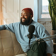 smiling bearded man gazing ahead while sitting on a couch in front of a microphone