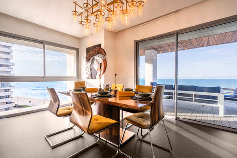 Salle à manger élégante avec chaises en cuir, table en bois et lustre design, offrant une vue imprenable sur la mer depuis la terrasse.