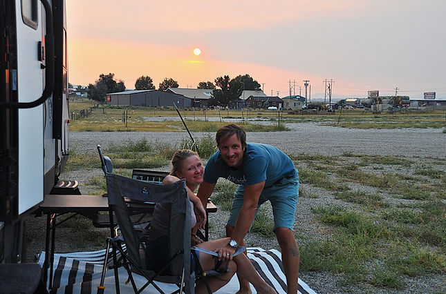 Alex and Liza of LAtravelFoxes, relaxing in camping chairs next to a travel trailer at a gravel boondocking campsite during sunset.