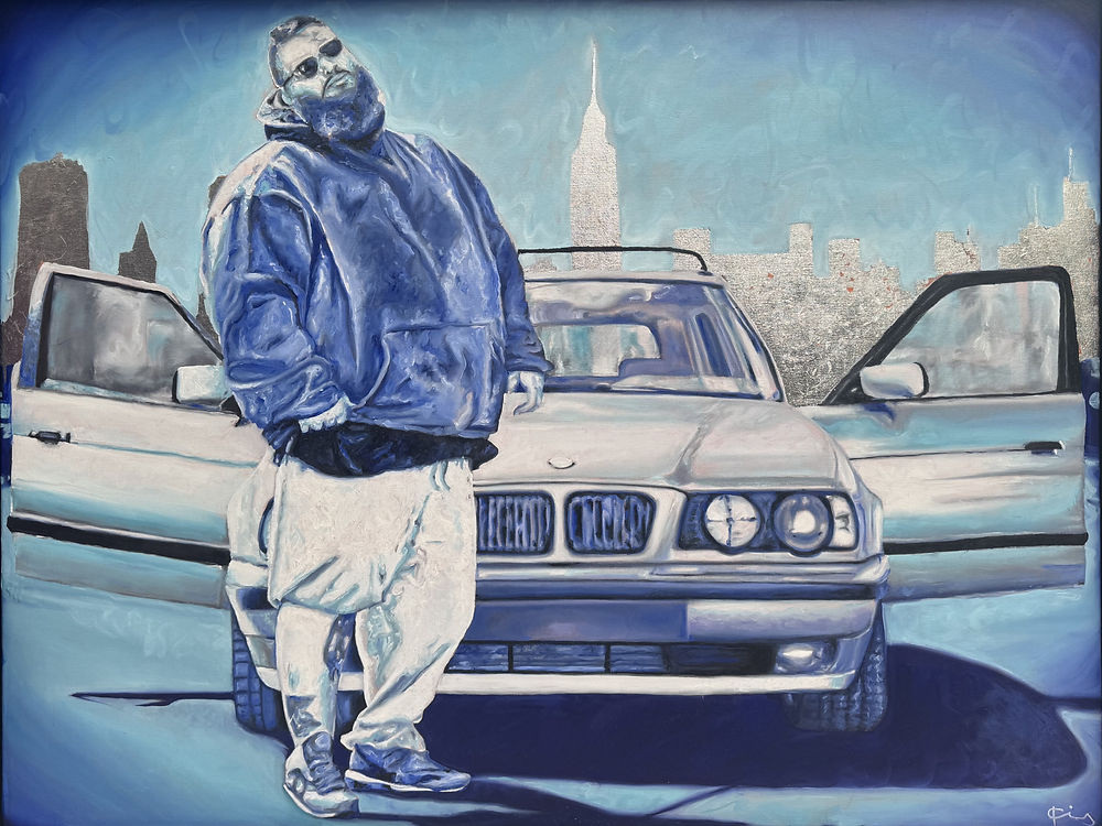 Bearded man (Action Bronson) in blues standing in front of a car with silver city skyline.
