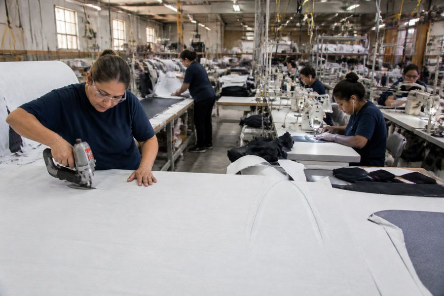Workers perform domestic contract sewing on an industrial factory floor with quality control stations in the background.