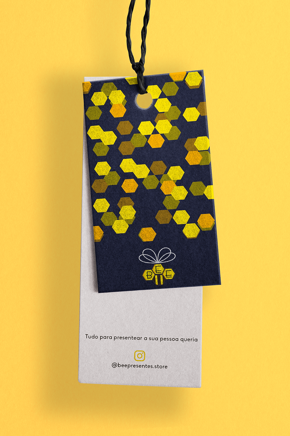 Uma etiqueta, com as cores azul e bege, em um fundo amarelo, a etiqueta azul está estampada com a logo Bee Presentes e possui um padrão de formas geométricas amarelas.