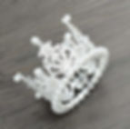 A small, silver-colored crown with intricate filigree details and pearls is placed on a wooden surface.