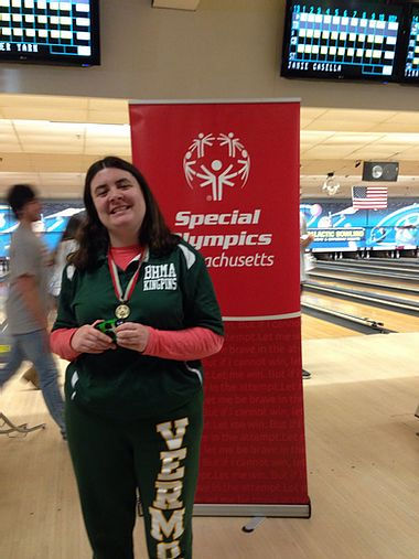 Image of Carly Golden, wearing her BHMA Kingpins green polo shirt, posing with her medal in front of the red Special Olympics pop-up sign at a bowling alley.