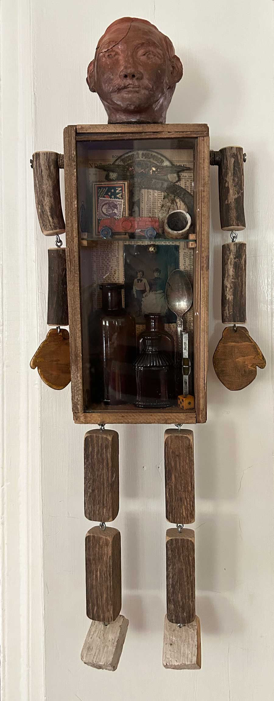 A sculptural assemblage. A man's shaped head, crude wooden arms and legs attached to a box, glass from, containing a number of objects a man of the 90th century might have used