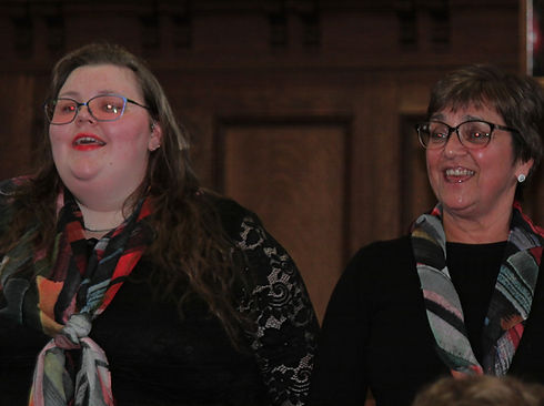 two women stand together and sing with smiles. They are wearing black with colourful scarves.