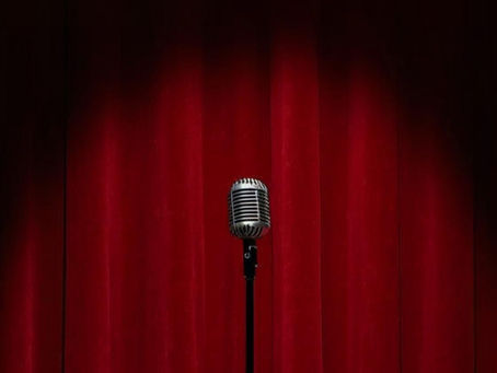 Das Bild zeigt eine Bühne mit einem roter Vorhang und ein Microfon, auf das ein Spotlight gerichtet ist.