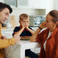 Das Bild zeigt eine Familie im Alltag. Mutter und Vater diskutieren und das Kind sitzt genervt daneben.