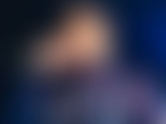 Homem jovem com barba e cabelo curto, escuro nas laterais e mais claro no topo, segura um microfone próximo à boca enquanto canta ou fala. Ele veste um moletom preto com estampa branca em letras grandes e estilizadas. A cena está iluminada por luzes fortes, com fundo escuro e algumas pessoas desfocadas ao fundo.