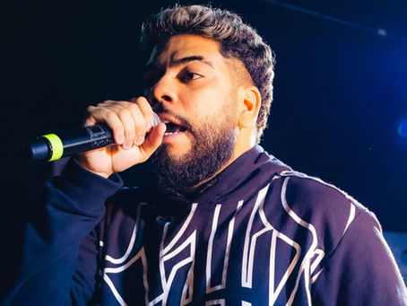 Homem jovem com barba e cabelo curto, escuro nas laterais e mais claro no topo, segura um microfone próximo à boca enquanto canta ou fala. Ele veste um moletom preto com estampa branca em letras grandes e estilizadas. A cena está iluminada por luzes fortes, com fundo escuro e algumas pessoas desfocadas ao fundo.