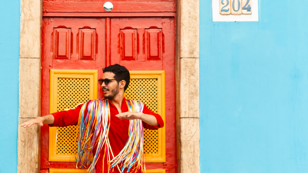 O cantor Gael Vila Nova posa em frente a uma porta vermelha com detalhes amarelos, em meio a uma parede azul vibrante. Ele veste uma camisa vermelha e um adereço de fitas coloridas nos ombros, remetendo à estética carnavalesca. Com óculos escuros, ele sorri e faz um movimento com os braços, transmitindo energia e leveza.
