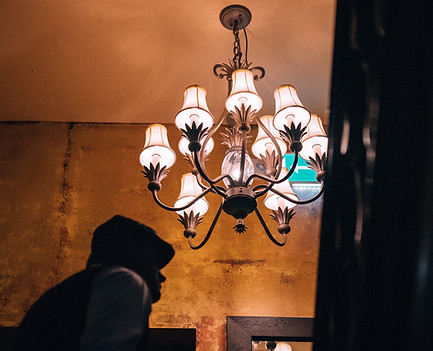 An upwards angle in a historic staircase. In the centre of the image is a large chandelier with sconce shades. The building has rustic pattenated yellow decorated walls. There is the silhouette of a man on the left of the image ahead of the photographer.