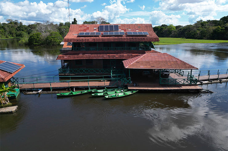 Pousada flutuante de telhado vermelho com painéis solares, barcos verdes atracados, em lago calmo sob céu azul.