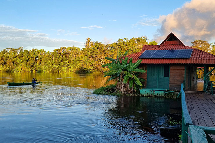Canoa com uma pessoa no rio ao lado de casa ribeirinha no Uakari Lodge, Reserva Mamirauá, com painéis solares, bananeira e céu dourado ao entardecer.