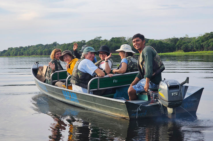 Grupo de turistas sorrindo e acenando em um barco pequeno no rio calmo do Uakari Lodge, Reserva Mamirauá, com floresta amazônica ao fundo.