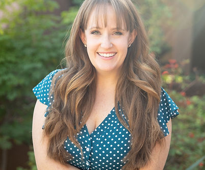 Sara Mersek. A caucasian woman in her 30s smiles joyfully at the camera. She has long brown hair that flows over both shoulders in curls. She is wearing a teal polka dot dress, and has one arm crossing her body holding her other arm. Behind her is lush greenery.