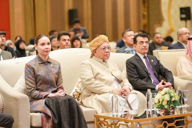 Formal audience at a conference: a woman, an elderly man in a gold turban, and a suited man sit on cream sofas in a wood-paneled hall