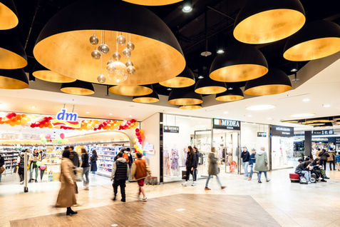 Blick in das Innere eines gut besuchten Shopping-Centers mit Fokus auf eine moderne Lampeninstallation an der Decke.
