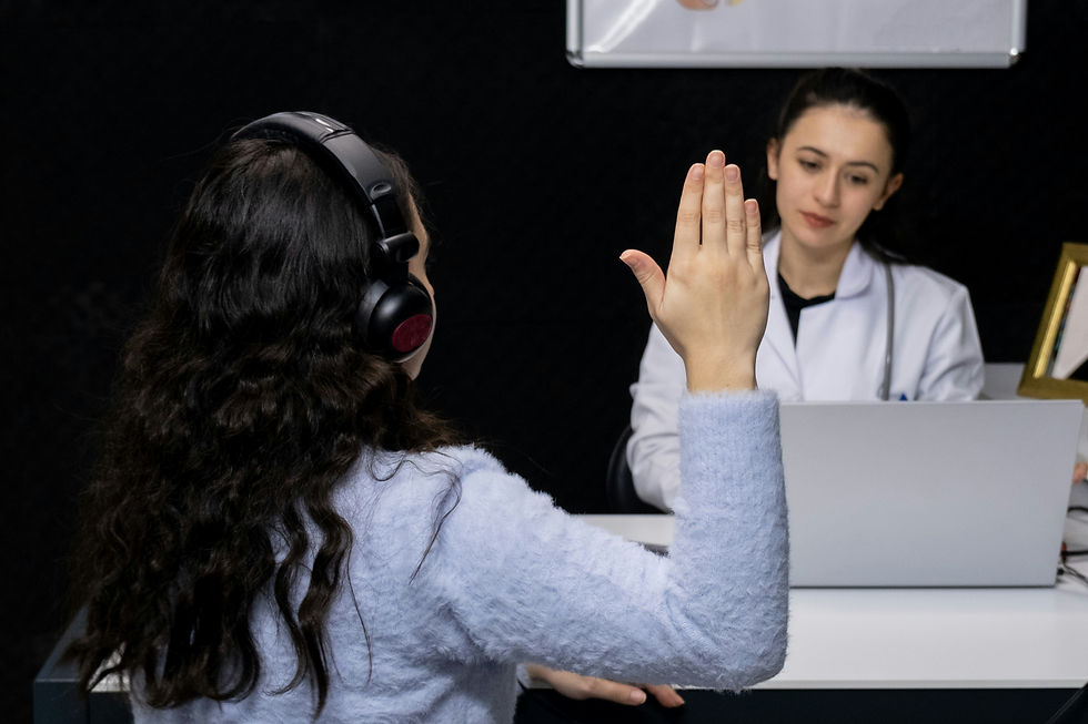 A woman with long, curly hair is undergoing a hearing test. She is wearing headphones and has her hand raised, indicating she hears a sound. In the background, a female audiologist, dressed in a white coat, is observing and taking notes on a laptop. The setting appears to be a medical office or clinic.