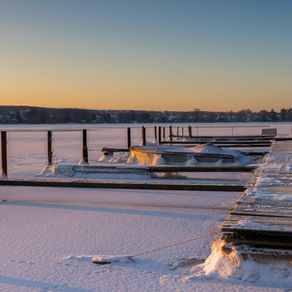 A snow-covered wooden dock extends over a frozen, snow-covered lake as the sun rises softly in the distance.