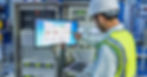 A worker in a hard hat and safety vest checks an industrial control screen in a plant control room while holding a laptop.