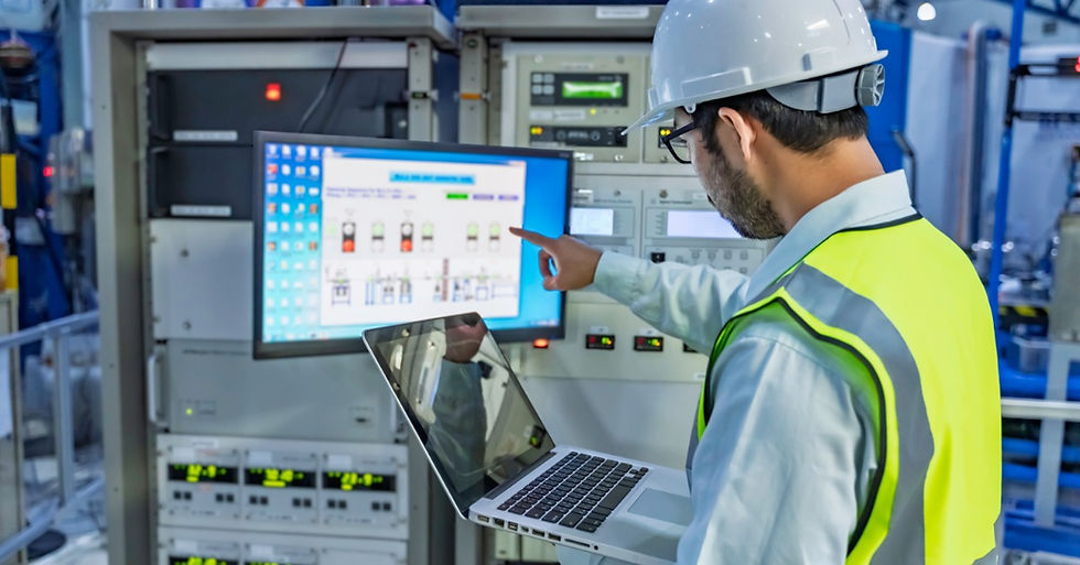 A worker in a hard hat and safety vest checks an industrial control screen in a plant control room while holding a laptop.