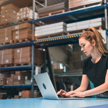 A woman in a warehouse surrounded by shelves with boxes on them wearing a black shirt while using a laptop.