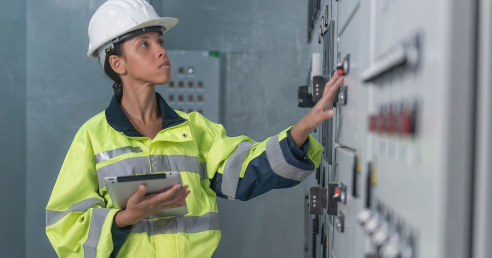 A female worker in a white hard hat and yellow safety jacket inspects a gray industrial control panel with a tablet.