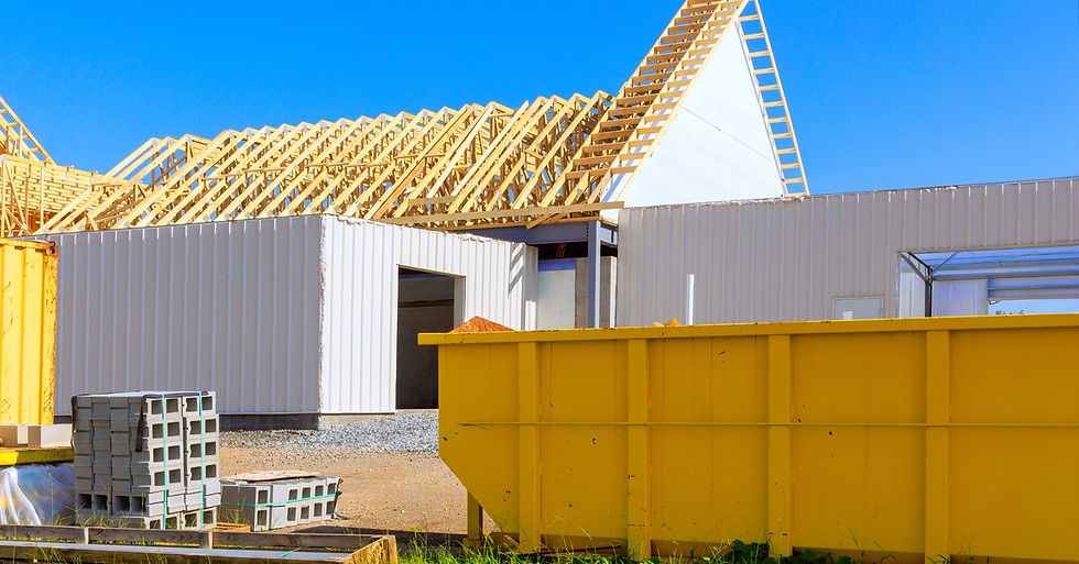A large yellow dumpster sits at a construction site next to building materials in front of a wooden building frame.