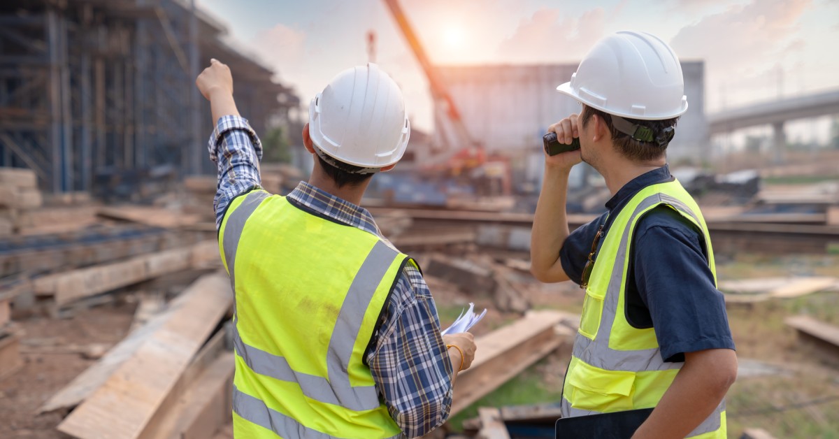 Two men are standing in a construction site. One is pointing and the other is on a radio.