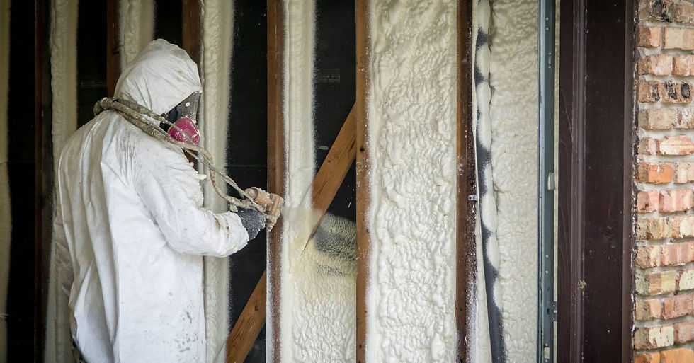 A worker wearing a full-body protective suit and mask using a spray gun to apply foam insulation into the walls of a building.