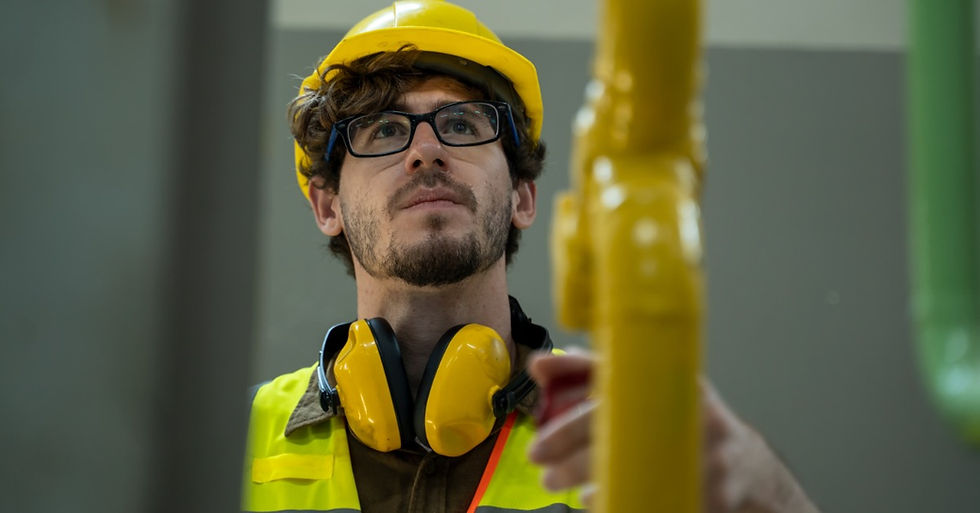 A worker in a yellow hard hat and safety vest inspects a bright yellow industrial pipe in an indoor facility.