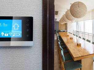 A black-and-silver smart thermostat with a blue screen mounted to a wall next to a room with a long table and chairs.
