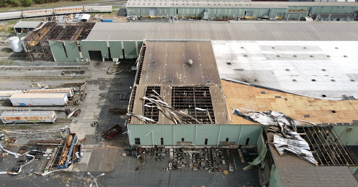 A large industrial building with part of the roof missing. The adjoining parking lot is full of debris.