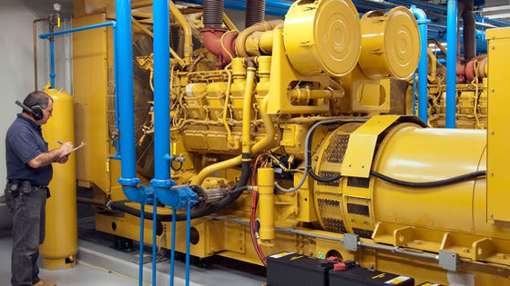 A man wearing jeans, protective ear gear, and a dark-colored polo is holding a clipboard next to an industrial generator.