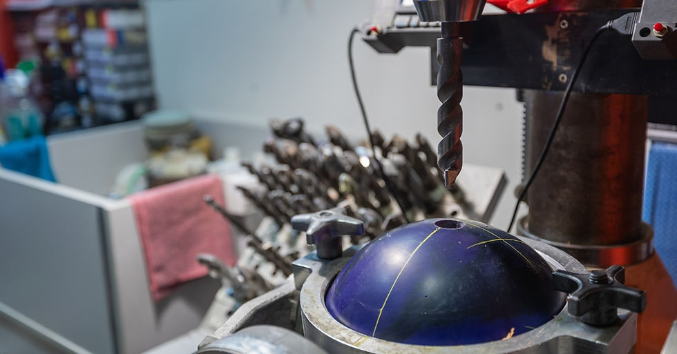 A blue and yellow bowling ball in a drill press, with the drill aligned over one of the ball's finger holes, in a workshop.