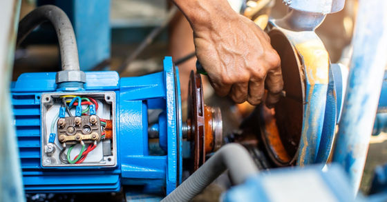 A close-up view shows a person's hand using a tool to turn a bolt on a blue industrial pump. The pump has hoses.