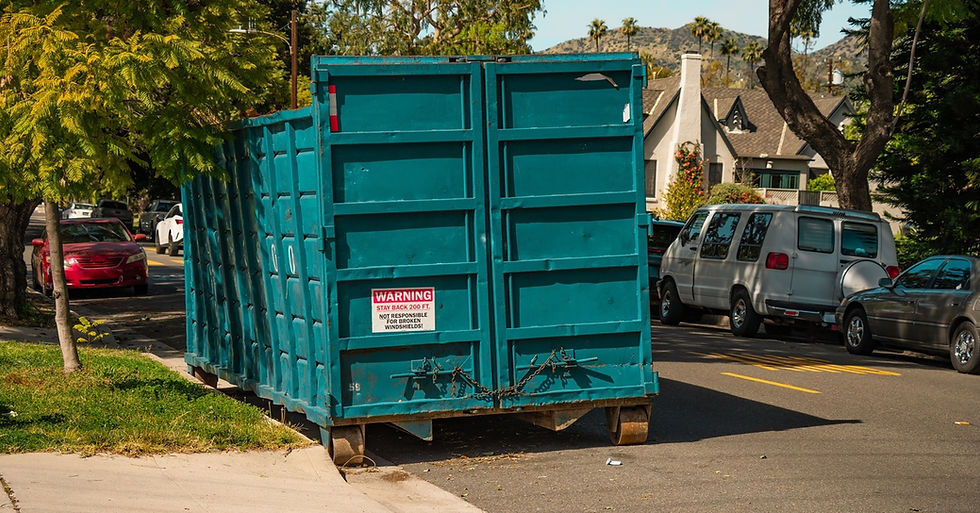 A large teal dumpster parked on the side of a road. It is situated in front of a driveway with trees next to it.