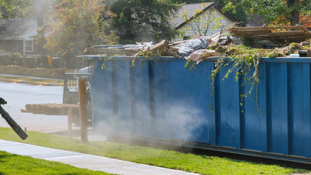 A blue roll-off dumpster is filled with landscaping debris in front of a yard where someone is using a leaf blower.