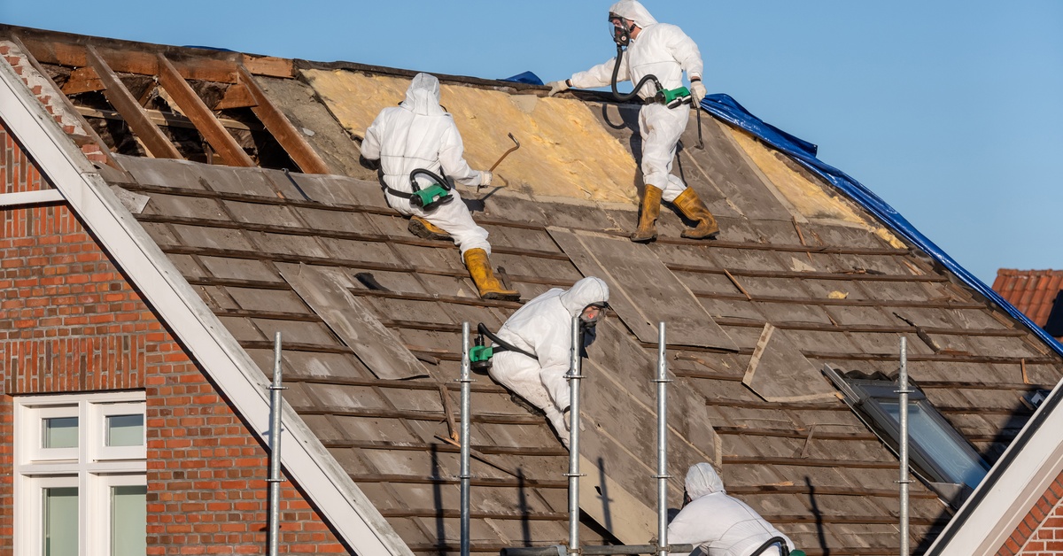 Several people are on a roof of a house. They are all wearing protective gear as they tear the roof apart.