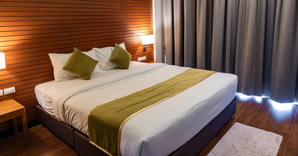 A hotel guestroom features brown wood paneling on the wall and gray curtains. The bed has white and green pillows.