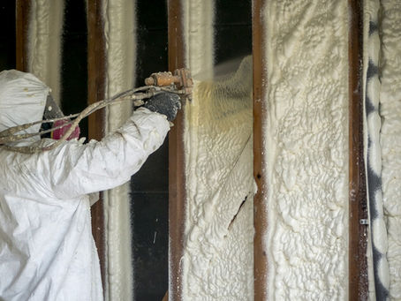 A person in a white full-body jumpsuit is spraying material against a wall. The material is insulation foam.