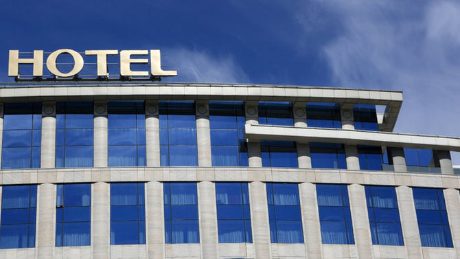 A large building stands with solid blue windows and the word "Hotel" on its roof. The sky in the background is blue.