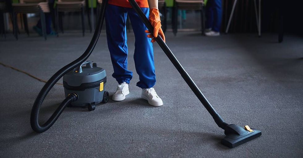 A person wearing blue pants, white shoes, and an orange shirt and gloves uses a vacuum to clean a gray carpet.