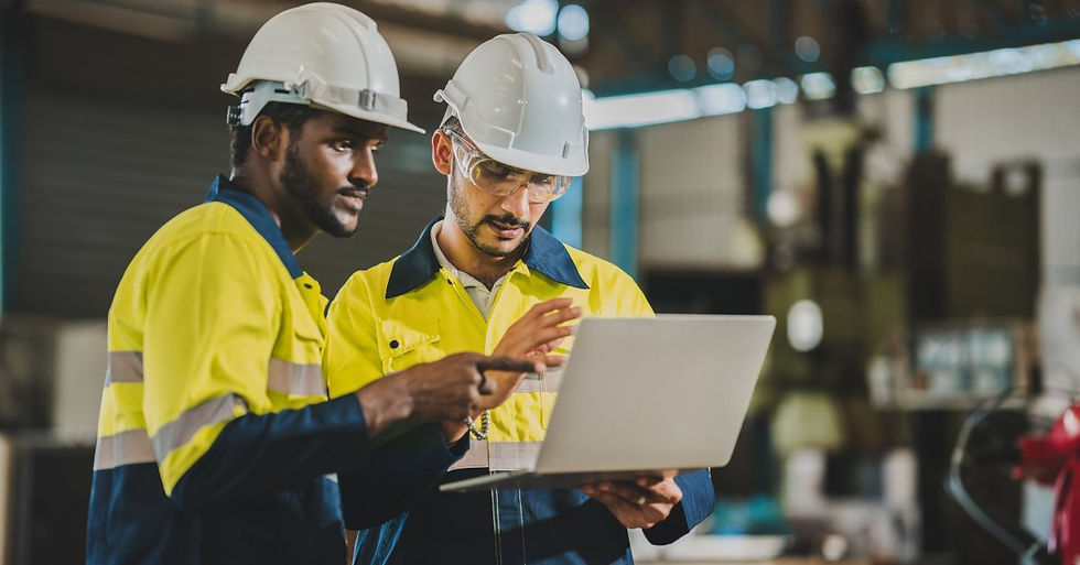 Two male workers in white hard hats and yellow and blue reflective jackets looking at a laptop while standing in a facility.