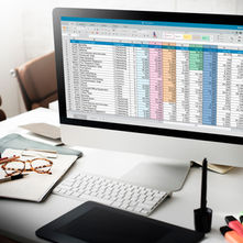 A desk with a clipboard and documents sitting in front of a computer with a spreadsheet program on the screen.
